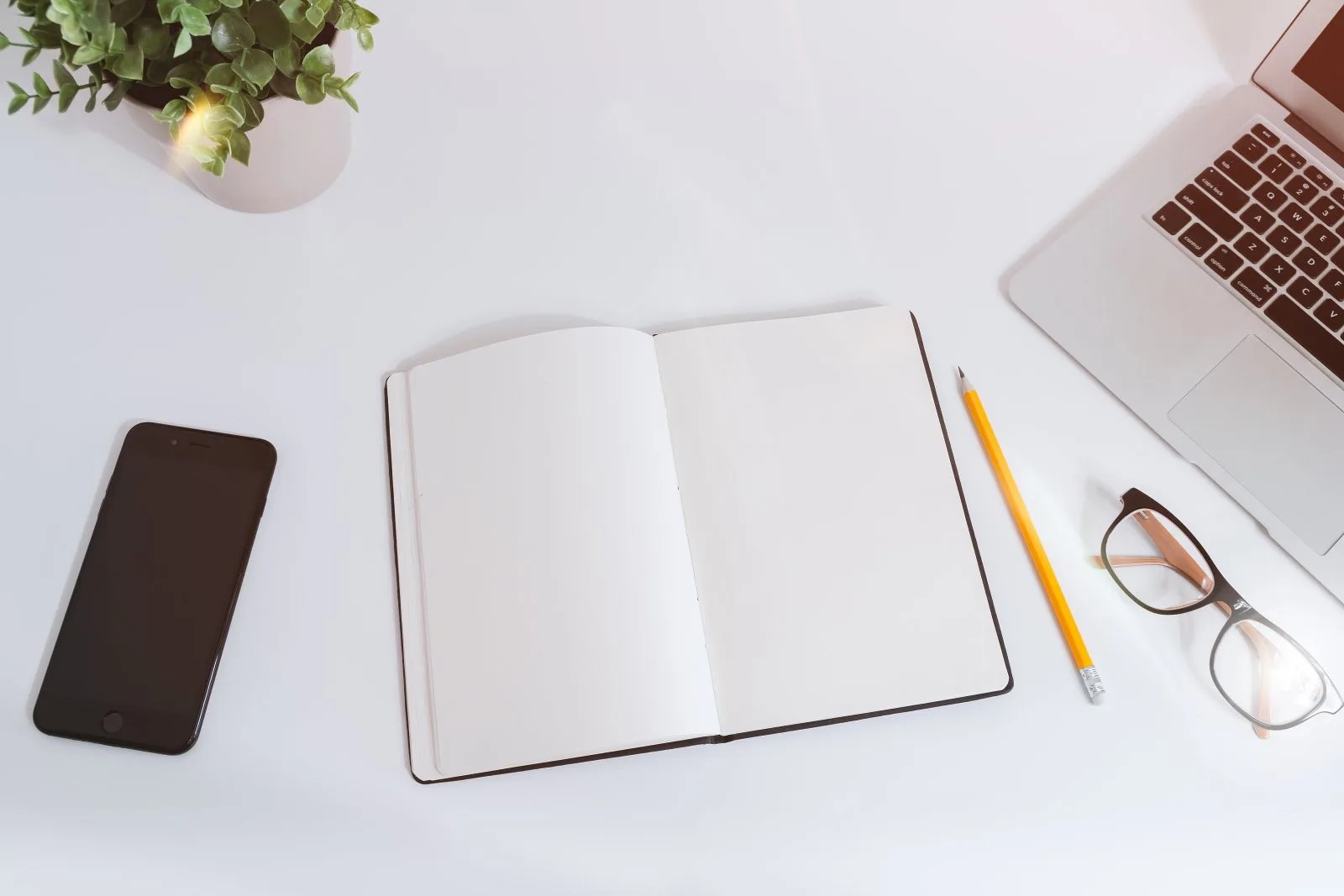 Mesa de trabajo blanca con un cuaderno abierto, un móvil, unas gafas y un portátil